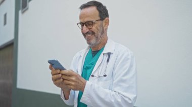 Güler yüzlü İspanyol erkek doktor hastane duvarının yanında steteskopla akıllı telefon kullanıyor ve gündüzleri gözlük takıyor.