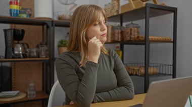 Masada, modern iç mekanda dizüstü bilgisayarla çalışan bir kadın, düşünceli ifadeli fikirler düşünüyor, günlük kıyafetler, arka planda dekore edilmiş raflar..