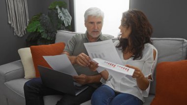 Orta yaşlı bir çift, oturma odasındaki finansmanı tartışıyor. Tablolar ve dizüstü bilgisayarla kanepe sergileniyor. Takım çalışması ve modern ev dekorasyonu..