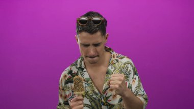 İspanyol asıllı genç adam pembe arka planda, çiçekli gömlekli ve modern tarzda neşeli yaz kaçışını yansıtan pembe elbisesiyle dondurma keyfine varıyor.