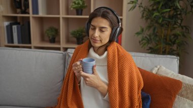 Evde dinlenen, kulaklık takan, elinde kupa tutan, turuncu battaniyeyle kaplanmış şık oturma odasında oturan, sükunetin, huzurun ve huzurlu ifadenin tadını çıkaran bir kadın..