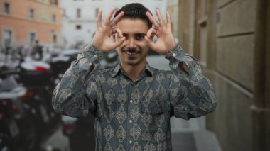 Latin kökenli genç adam şehir manzarası ile çevrili bir şehir caddesinde el hareketi yapıyor..