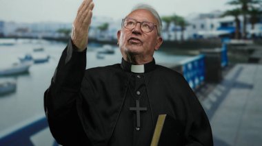 Siyah cüppeli kıdemli rahip, güneşli bir sahil gezisinde mavi korkuluklar ve görünür teknelerle düşünceli bir şekilde el kol hareketi yapar..