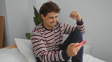 Genç adam rahat bir yatak odasında akıllı bir telefon kullanarak evde mutluluk ve rahatlık göstererek kutlama yapıyor..