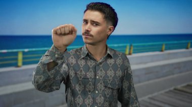 Deniz kenarında gezinen genç İspanyol bir adam berrak mavi bir gökyüzü ve okyanus arkaplanına karşı derin bir ifadeyle, sakin bir açık deniz manzarası sergiliyor..