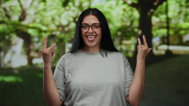 Parkta gözlüklü bir kadın güneşli bir günde gülümseyen gri tişörtlü, güveni ve güveni olan ağaçlarla dışarıda taş jestler yapıyor..