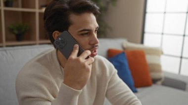 Modern oturma odasında oturan İspanyol asıllı genç adam rahat ve düşünceli, çağdaş iç mekan tarzını ve sakin atmosferi yakalıyor..