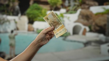 Arka planda bir adamın olduğu bir tatil havuzu kenarında Norveçli bir kronun elini tutmak, bir tatil ortamında seyahat ve finans temasını vurgulamak.