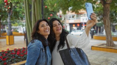 Şehir parkında selfie çeken iki gülümseyen kadın şehir hayatı ve yeşillik arasında neşeli birliktelik ve dostluğu yakalıyorlar..