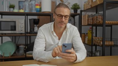 Modern ofis ortamında akıllı telefon kullanan gözlüklü orta yaşlı bir adam, çeşitli mutfak malzemeleriyle çevrili raflarla çevrili, sıradan bir çalışma ortamını ima ediyor..
