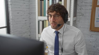 Gözlüklü ve kulaklık takan genç bir adam modern bir ofiste çalışıyor. Etrafı raflarla çevrili ve beyaz tuğladan bir duvarla çevrili. Üretim ve uzmanlık öneriyor..