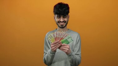 Norveç kronunu izole edilmiş turuncu arka planda tutan adam gülümsüyor, zenginlik ve mutluluk sergiliyor..