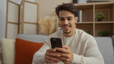 Genç adam rahat bir oturma odasında akıllı telefon kullanırken gülümsüyor, rahat ve modern bir iç dekorasyon sergiliyor..