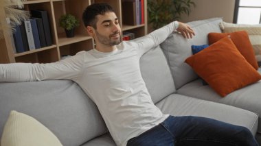 Genç yakışıklı bir adam şık bir oturma odasında rahat bir koltukta dinleniyor, modern bir ev ortamında huzur ve rahatlık hissi yayıyor..