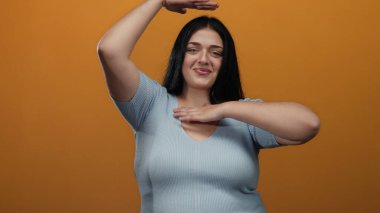 Parlak turuncu arka planda elleri ile gülümseyen kadın oyuncu bir şekilde kıvrımlı figürler sergiliyor..