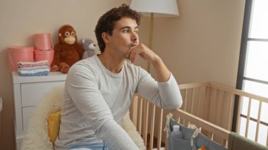 Genç adam, yumuşak oyuncaklar ve pastel dekorlarla çevrili bir bebeğin beşiğinin yanındaki rahat bir yatak odasında düşünceli bir şekilde oturuyor ve evde huzurlu bir iç atmosfer yaratıyor..