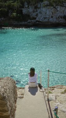 Güneşli bir günde Mallorca 'da turkuaz suları olan Cala Lombars plajının keyfini çıkaran kadın, Akdeniz sahilinin sükunetini ve doğal güzelliğini vurguluyor..