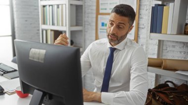 Ofis ortamında masa başında çalışan yakışıklı, beyaz gömlek ve kravat takan, modern iş yerinde bilgisayar ekranı ile meşgul olan, profesyonel çevreyi gözler önüne seren yakışıklı bir İspanyol..