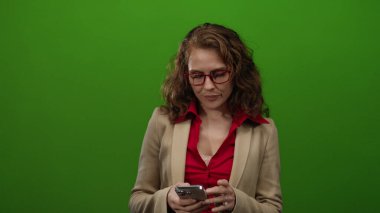 Akıllı telefon kullanan bir kadın, izole edilmiş yeşil arka planda kırmızı bluz ve kırmızı gözlüklü bej ceket giyiyor. Stüdyoda teknoloji ve iletişim kavramlarını tasvir ediyor..