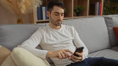 Genç adam oturma odasında akıllı telefon kullanıyor, koltukta dinleniyor, rahat iç dekorasyonla çevrili, modern yaşam tarzını ve teknolojik bağlantılarını gösteriyor..