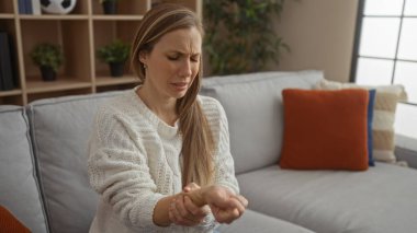 Rahat bir oturma odasında, modern bir koltukta bilek acısı çeken bir kadın, günlük bir kazak giyiyor, dekoratif yastıklarla ev ortamında rahatsızlığını dile getiriyor..
