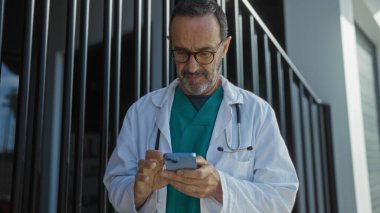 Gündüz vakti şehir sokaklarındaki hastanenin önünde telefon kullanan doktor ceketli, olgun İspanyol bir adam.