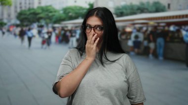 Gözlüklü genç bir kadın marketin dışındaki işlek şehir sokağında dikiliyor. Elleriyle ağzını kapayarak şehrin bulanık arka planına şaşırıyor..