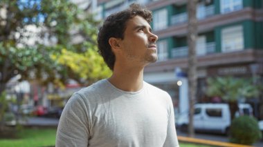 Şehir arka planında hayat dolu bir şehir parkına özenle bakan genç bir adam ve açık hava kentsel deneyimini geliştiren yemyeşil bir yer.