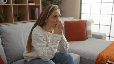 Sıcak bir odada rahat bir kanepede oturan kadın düşünceli görünüyor, sıcak dekor ve pencereden süzülen doğal ışıkla çevrili..