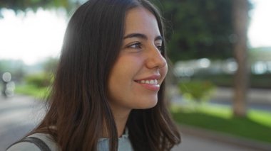 Şehir parkında gülümseyen bir kadın, yemyeşil ve güneş ışığıyla doğal güzelliğini ve esmer saçlarını ön plana çıkaran neşeli ve barışçıl bir deneyim öneriyor..