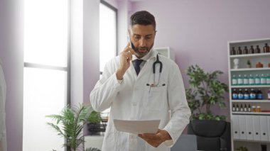 Doktor, hastane bürosunda dokümanları incelerken telefonla konuşuyor. Etrafı ilaç şişeleri ve bitkilerle çevrili. Parlak ışık altında..