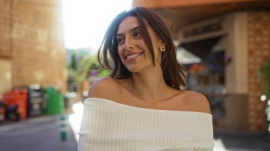 Kentsel bir alanda gülümseyen bir kadın, canlı bir şehir zeminine karşı özgüven ve güzellik sergiliyor. Güneş ışığı esmer saçlarını ve omuzlarını aydınlatıyor..