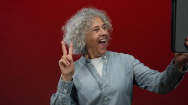 Gri saçlı bir kadın canlı kırmızı bir duvara karşı selfie çekerken barış işareti yapıyor. Eğlenceli ve canlı bir atmosferde rahat bir gömlek giyiyor.