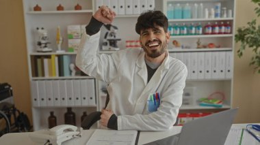 Klinikte gülümseyen genç bir adam laboratuvar önlüğü giyiyor ve ofis malzemeleriyle dolu bir masanın arkasında iyi organize edilmiş bir iş ortamında kutlama yapıyor.