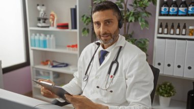 Klinikteki adam kulaklık takıyor ve arka planda tıbbi malzemeler ve bitkilerle dolu raflarla dolu modern bir hastane odasında pano tutuyor..