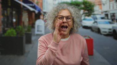 Şehir sokaklarında gülümseyen ve konuşan kır saçlı kadın, hareketli bir şehir ortamında olgun bir özgüvenin ve açık hava aktivitesinin özünü yakalıyor..