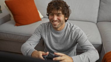 Yakışıklı adam kanepede video oyunları oynarken gülümsüyor rahat bir oturma odasında rahat turuncu yastıklar ve sıcak bir ortam ile..