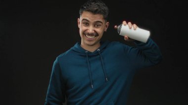 Siyah arka planda sprey boya kutusu tutan genç bir adam, mavi kapüşon giyiyor, stüdyo ortamında neşeli duygular ifade ediyor..