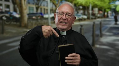 Başrahip bir şehir caddesinde dururken bir kitabı işaret ediyor. Çevresi kentsel unsurlar ve açık hava ortamında yeşilliklerle çevrili..