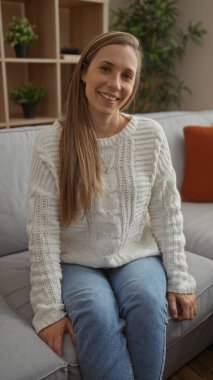 Evde rahat bir dairede rahat bir şekilde gülümseyen bir kadın rahat bir süveter ve arka planda bitkiler olan kanepede oturan bir kot pantolon giyiyor.