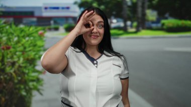 Uzun saçlı genç bir kadın gözünün üstüne iyi bir jest yapıyor, yeşil bir sokakta gülümsüyor ve geçmişi bulanık..