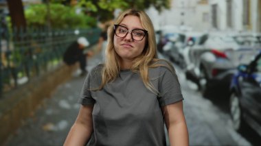 Gri tişörtlü ve gözlüklü bir kadın park etmiş arabaları ve arka planda yeşilliği olan bir şehir caddesinde dikiliyor düşünceli duygularını ifade ediyor..