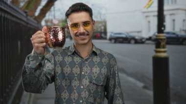 Şablon gömlekli ve sarı gözlüklü genç İspanyol bir adam şehir sokaklarında bir bakır bardak tutuyor, açık şehir ortamında neşeli bir hava yayıyor..