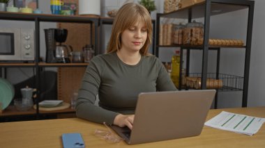 Ofis mutfağında dizüstü bilgisayarda çalışan bir kadın. Raflar ve evrak işleri yakınlarda. Üretken iş ortamını somutlaştırıyor..