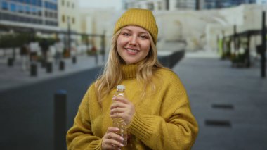 Sarı bir süveter giymiş, elinde su şişesi olan bir kadın şehir sokaklarında, gün içinde sakin ve neşeli bir hava iletiyor..