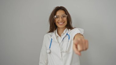 Beyaz önlüklü, steteskoplu, kendinden emin bir şekilde gülümseyen kadın doktor, izole edilmiş beyaz arka planı işaret ediyor..
