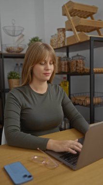 Ofiste dizüstü bilgisayarla çalışan bir kadın. Etrafı raf ve masa eşyalarıyla çevrili. Odaklanmış ve üretken bir çalışma alanı ortamı yaratıyor..