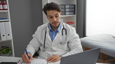 Steteskoplu İspanyol erkek doktor, yoğun bir gün boyunca tıbbi ekipmanlarla çevrili bir klinikte çalışıyor, dokümanlar ve laptoplar üzerinde yoğunlaşıyor..