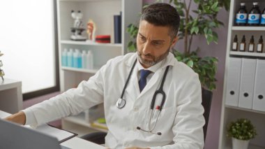 Klinikte çalışan beyaz önlüklü doktor etraftaki tıbbi ekipman ve çizelgelerle bilgisayar ekranına odaklanıyor..