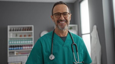 Yakışıklı İspanyol erkek doktor iyi ışıklandırılmış bir klinikte sıcak bir şekilde gülümsüyor. Deniz mavisi önlük ve steteskop giyip profesyonel ve misafirperver bir sağlık hizmeti sunuyor..